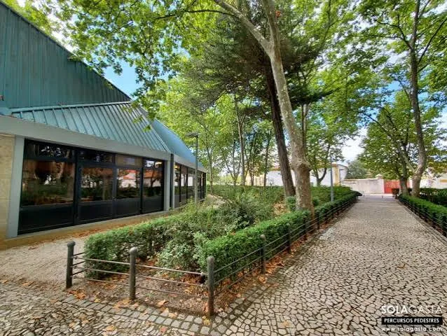 Tree-shaded paved path beside the glass-fronted Os Cucos restaurant